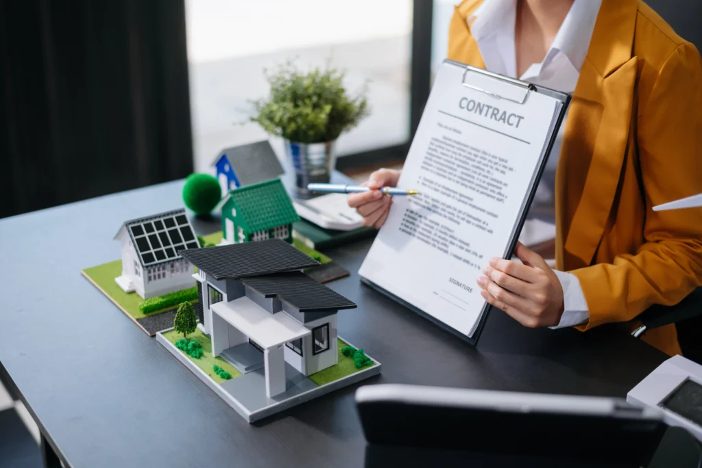 A person in a yellow blazer holding a contract while reviewing it with a model of houses, including a solar panel house and a green plant, on a desk.