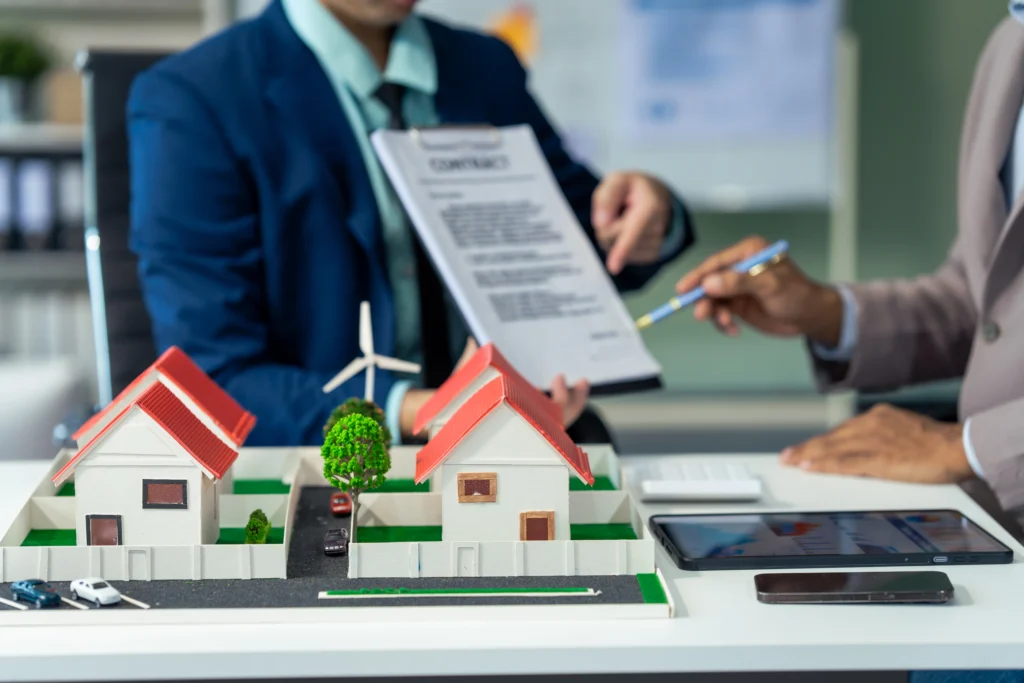 Two professionals in suits discussing a contract over a desk with a miniature house model, a tablet, and a smartphone, in a modern office setting.