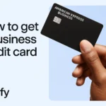 A close-up of a hand holding an American Express business credit card against a blue background, with the text "How to get a business credit card" and the word "Zavify" on the left.