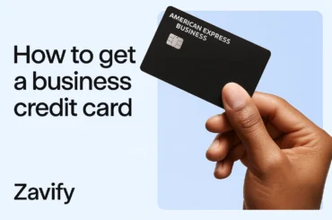 A close-up of a hand holding an American Express business credit card against a blue background, with the text "How to get a business credit card" and the word "Zavify" on the left.